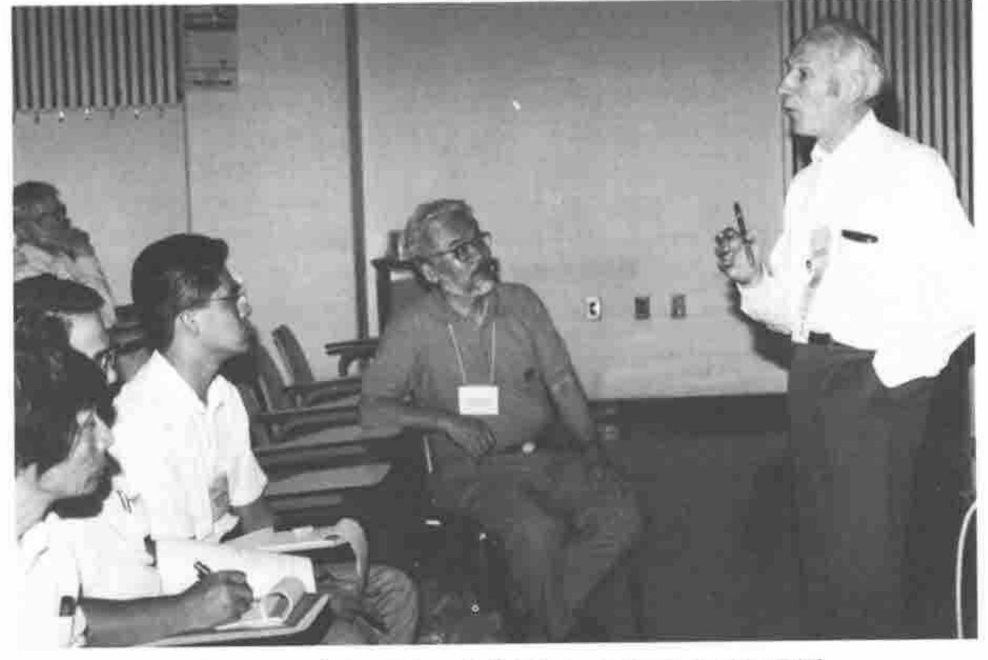 Donald McIntyre, standing, addressing a seated group taking notes