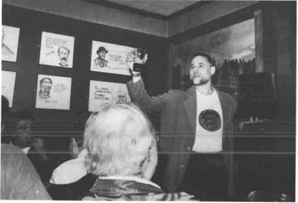 A man with a raised glass addressing a room hung with caricatures