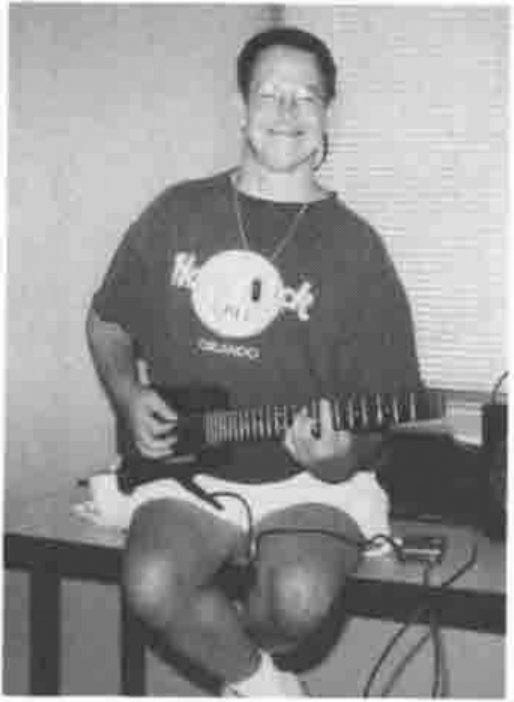 Alan Graham playing an electric guitar, seated on a table