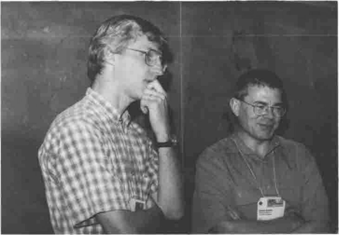 Two men talking: one with hand to chin, the other smiling with arms folded