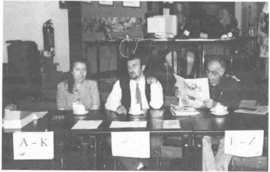 Photograph: three people seated at the registration desk, with alphabetical signs for registration