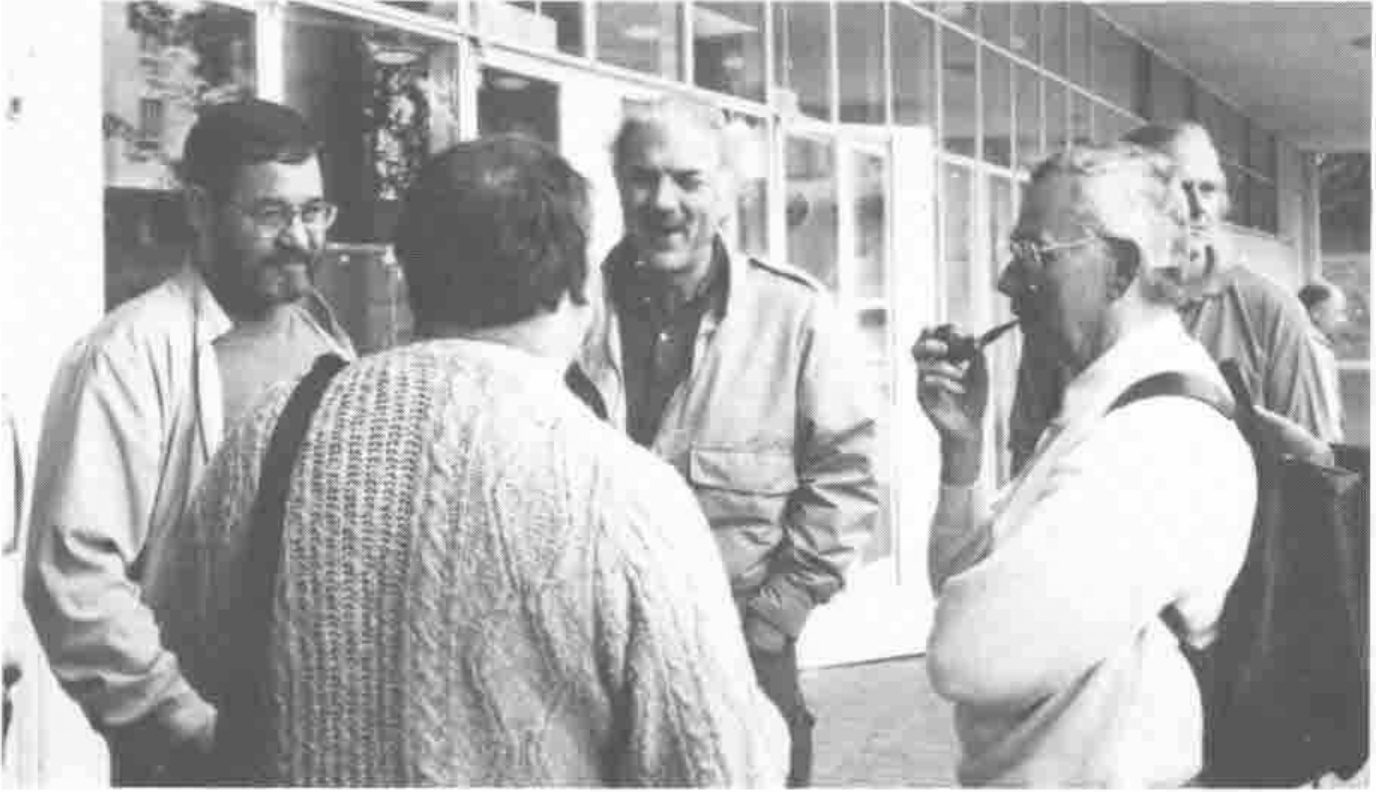Photograph of five delegates talking outside a glass-fronted building; one, smoking a pipe, wears a rucksack