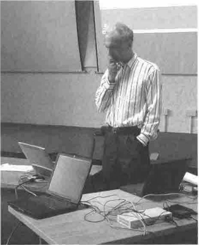 A man in a striped shirt, hand to his chin, standing behind a table with two laptops and their cables