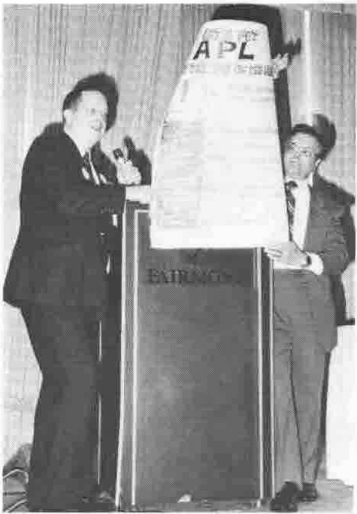 A man speaking into a microphone at a lectern, beside a tall paper cone marked APL held by another man