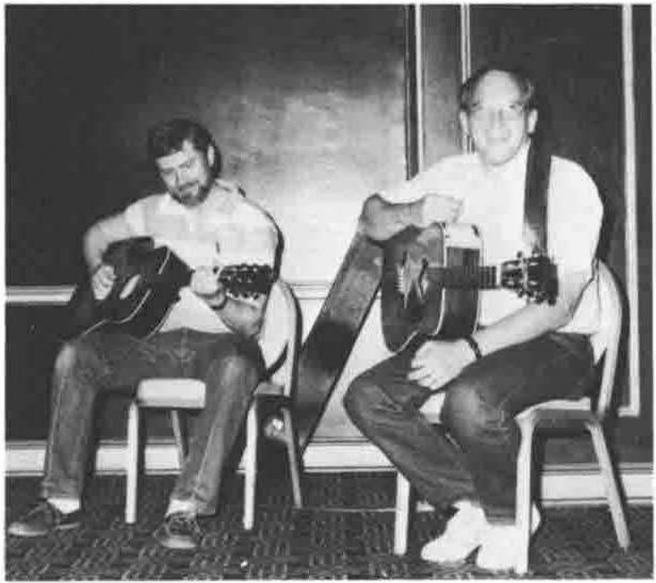 Two men seated on chairs on a stage playing acoustic guitars