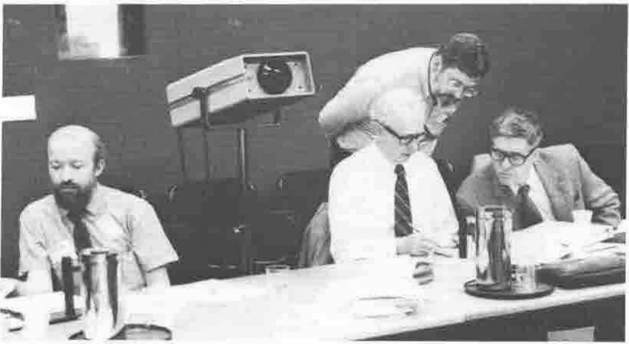 Four men at a table, papers in front of them, beside an overhead projector