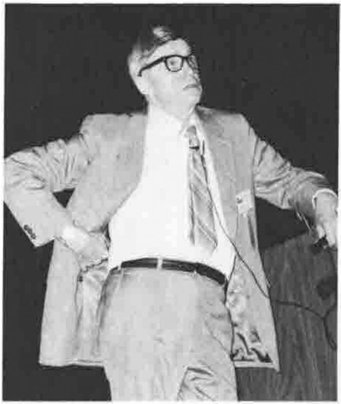 A man in glasses, jacket and tie, standing with one hand on his hip and looking upwards