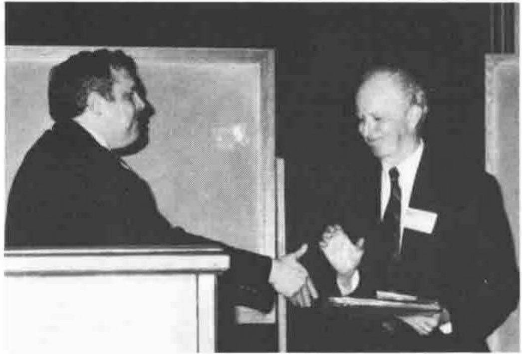 A man at a lectern shaking hands with an older man who holds a plaque