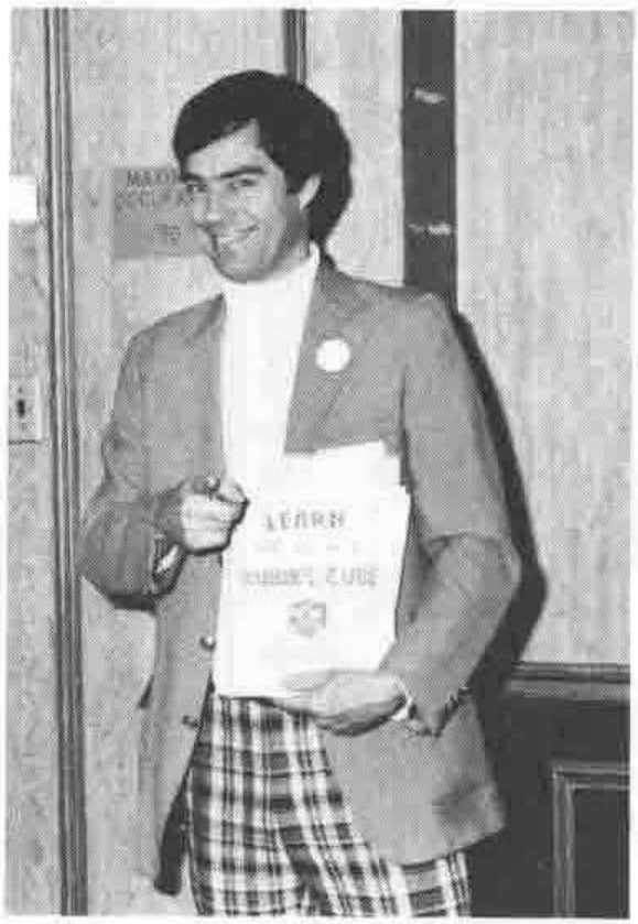 A smiling man in a jacket and checked trousers holding a booklet headed LEARN and pointing at the camera