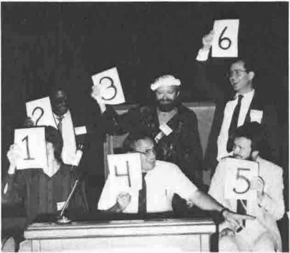 Six people on a stage holding up cards numbered 1 to 6, some of the cards hiding their faces