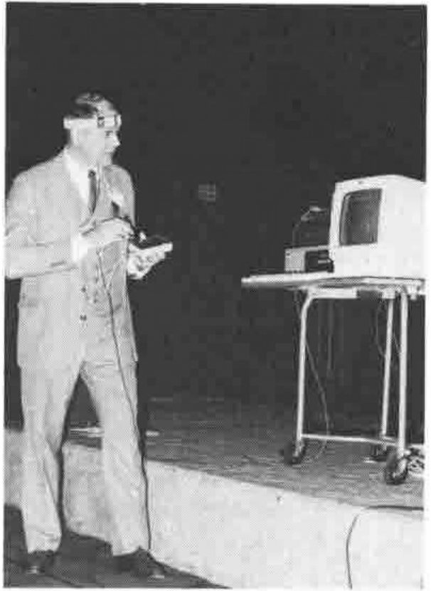 A man on a stage wearing a headband with electrodes, holding a box wired to it, near a computer on a trolley