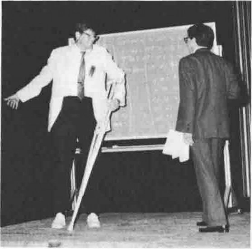 A man in a light jacket on crutches gesturing on a stage, while another man with papers under his arm faces a flip chart