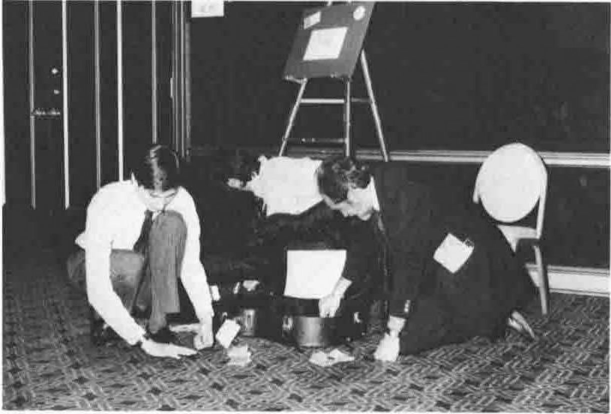 Two men crouching on a carpeted floor, picking up small objects spilled beside an open case, in front of an easel