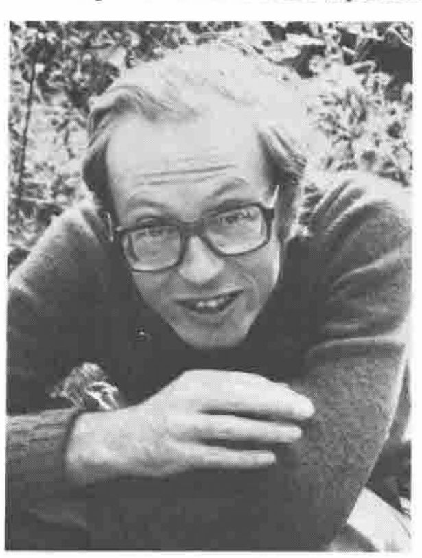 Adrian Smith, in glasses and a sweater, leaning on his folded arms in front of foliage