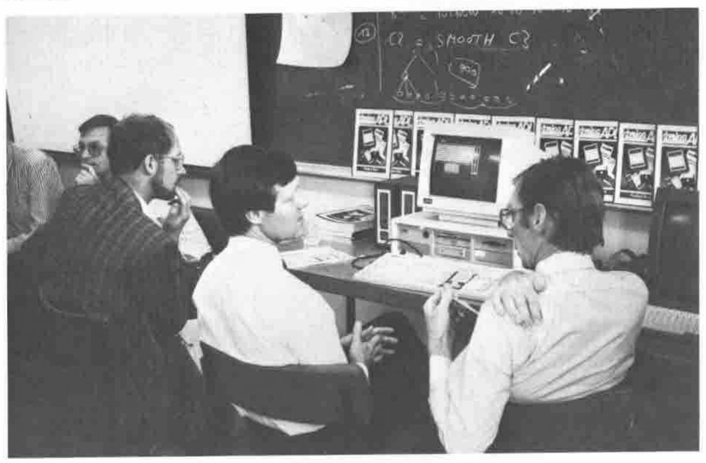 Photograph: four men at a demonstration table; one, seated at a PC, turns to talk to another beside him, with a row of Dyalog APL boxes along the wall and a blackboard behind