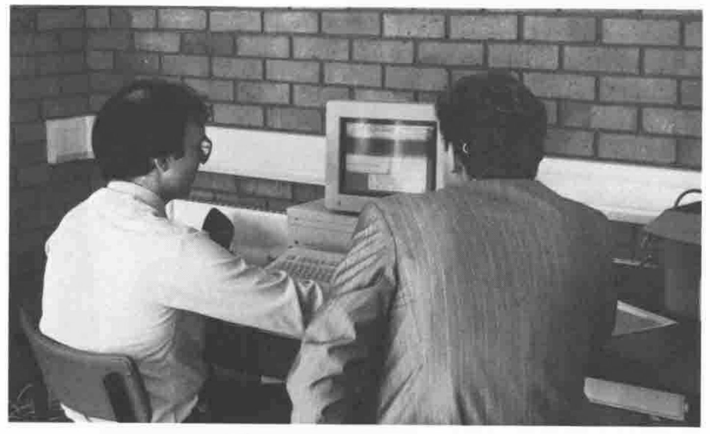 Photograph: two men, seen from behind, seated at a computer against a brick wall