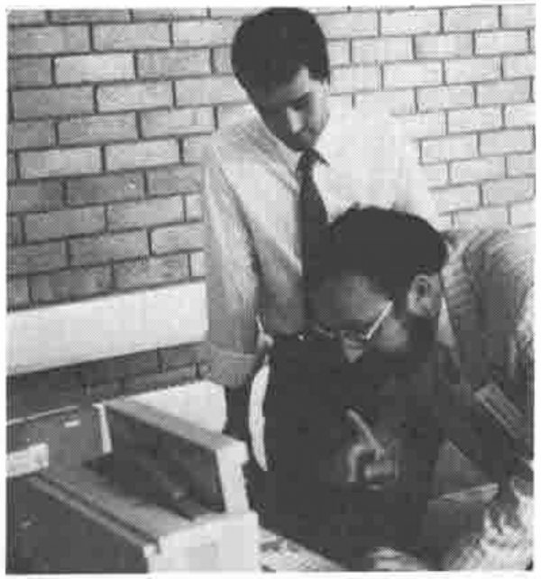 Photograph: a young man in shirt and tie stands looking down over the shoulder of a bearded man in glasses, seated at a computer against a brick wall
