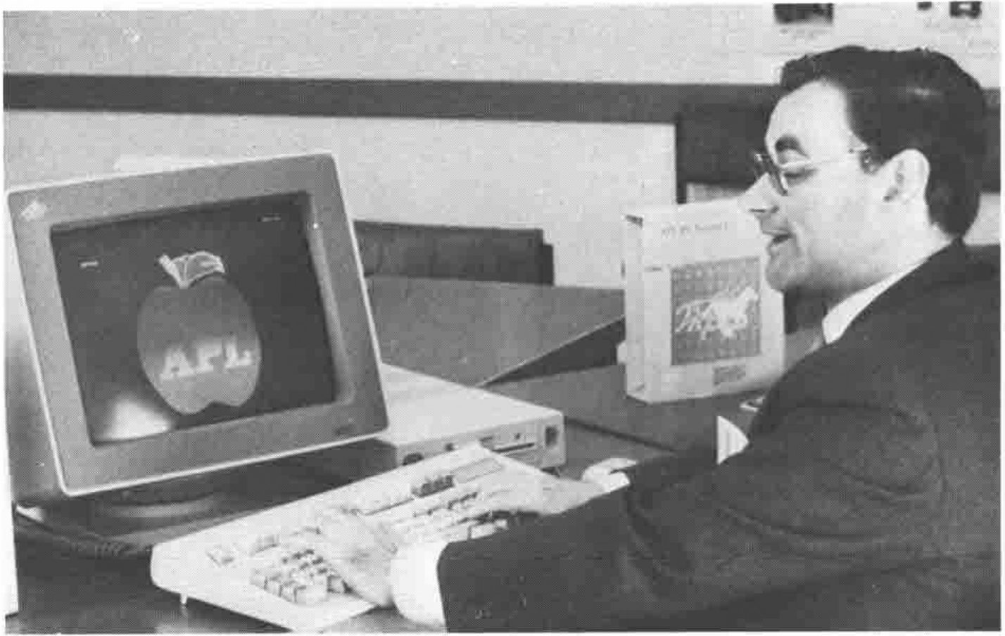 Photograph: a man in glasses and a dark suit types at an IBM PS/2 keyboard; the monitor shows an apple with “APL” across it, and an APL2/PC product box stands beside him