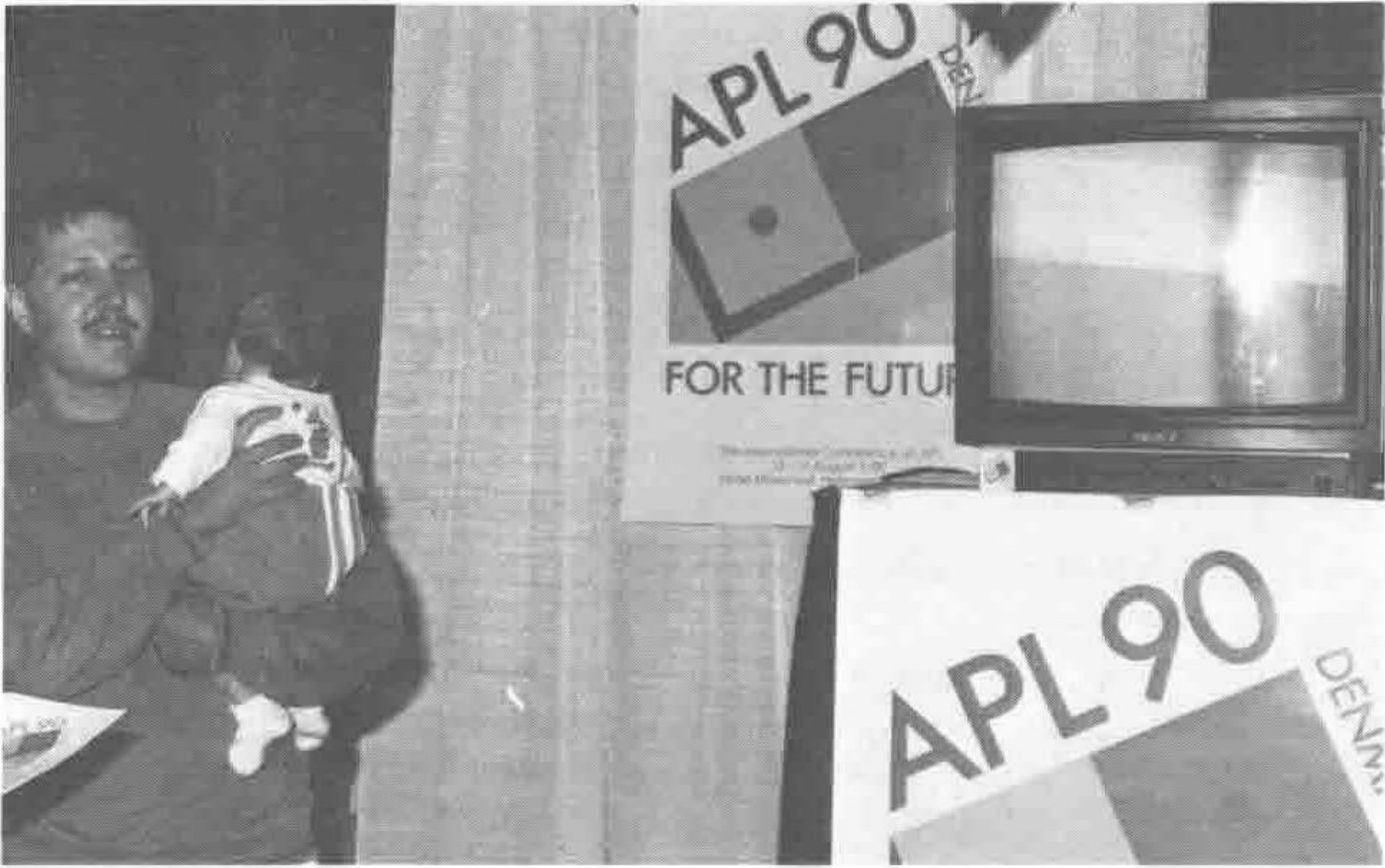 Photograph of a man holding a baby beside APL90 Denmark posters (“APL90 … For the Future”) and a television monitor