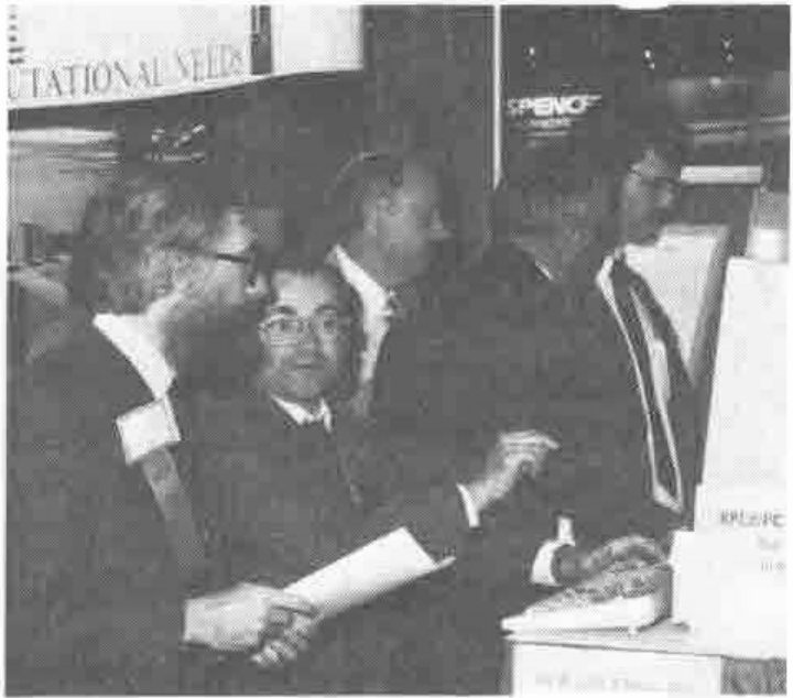 Photograph of a group of men gathered round a seated man at a PC keyboard on the APL2/PC stand