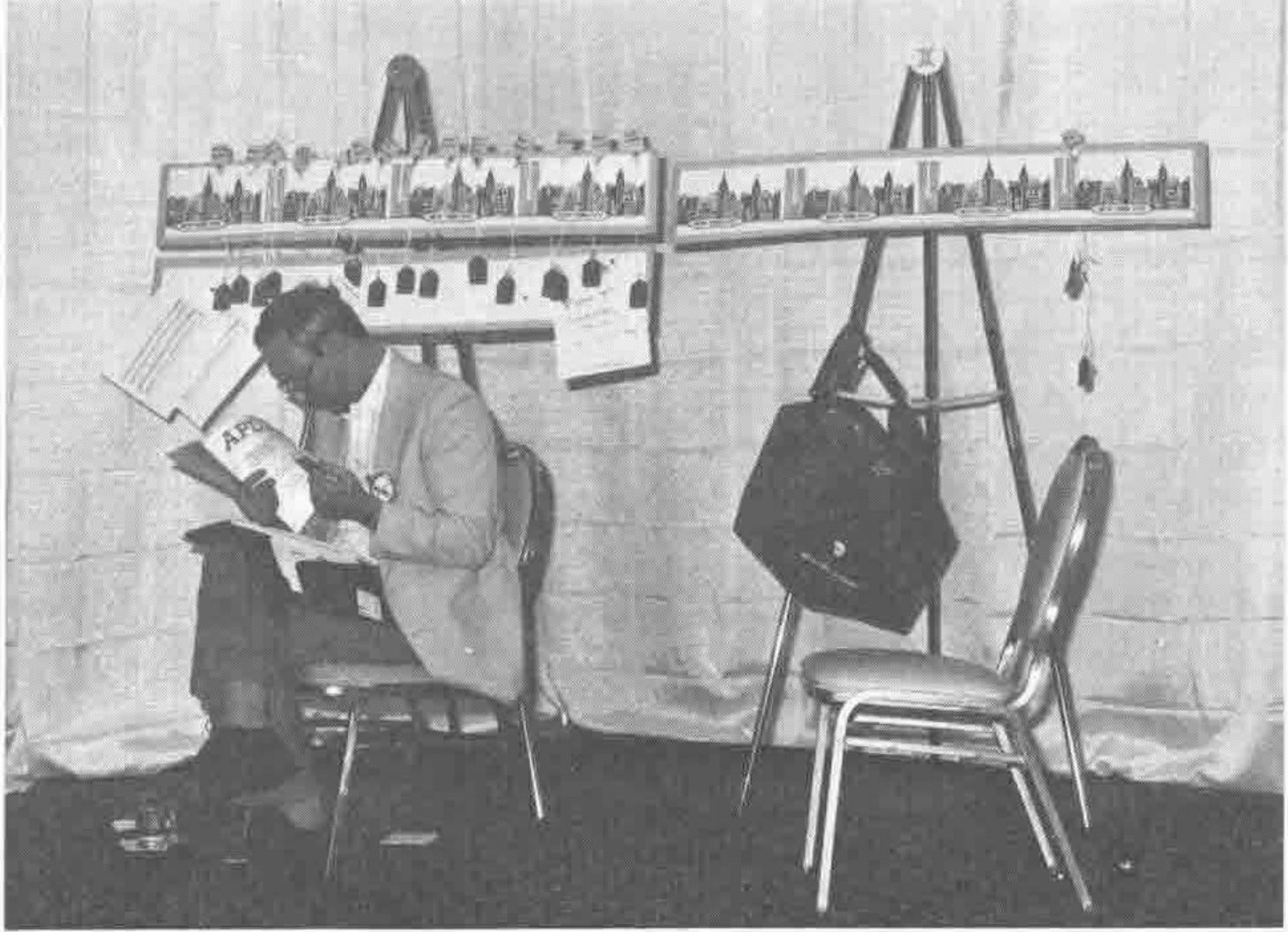 Photograph of a man sitting on a chair reading an APL book in front of easels hung with New York skyline pictures