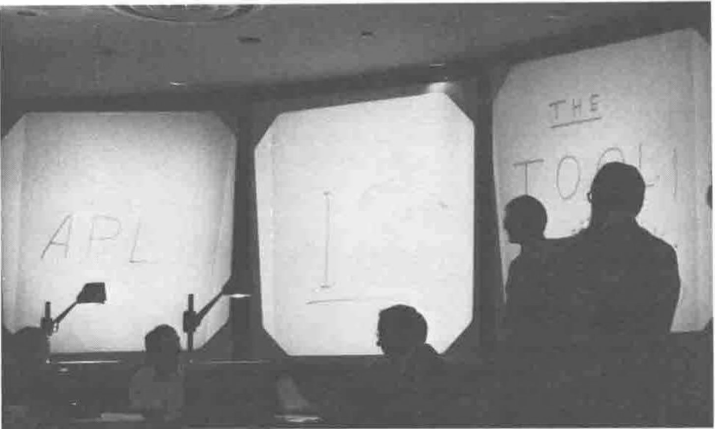 Photograph of three projection screens in a darkened hall, with people in silhouette at the overhead projectors; the screens show “APL” and “THE TOOL”