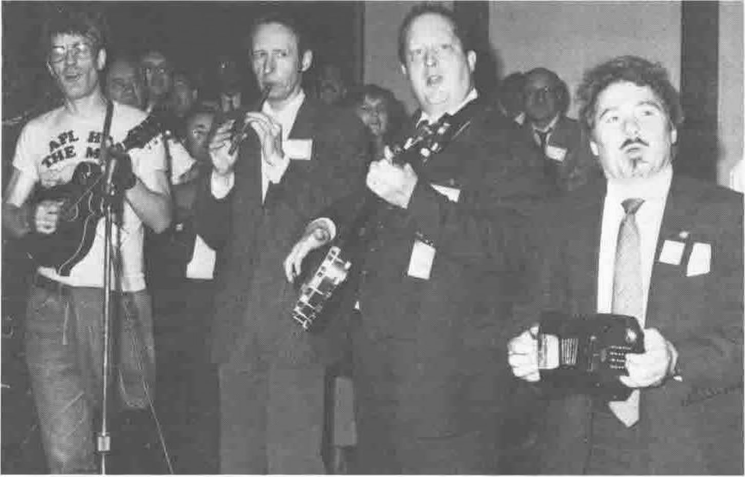 Photograph of four musicians playing: a man in an APL T-shirt with a mandolin, a whistle player, a man with a tambourine, and a concertina player
