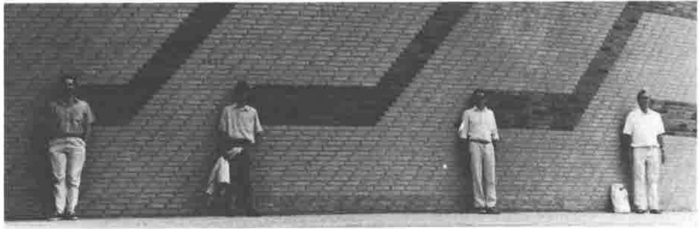 Four men standing apart from one another against a long brick wall