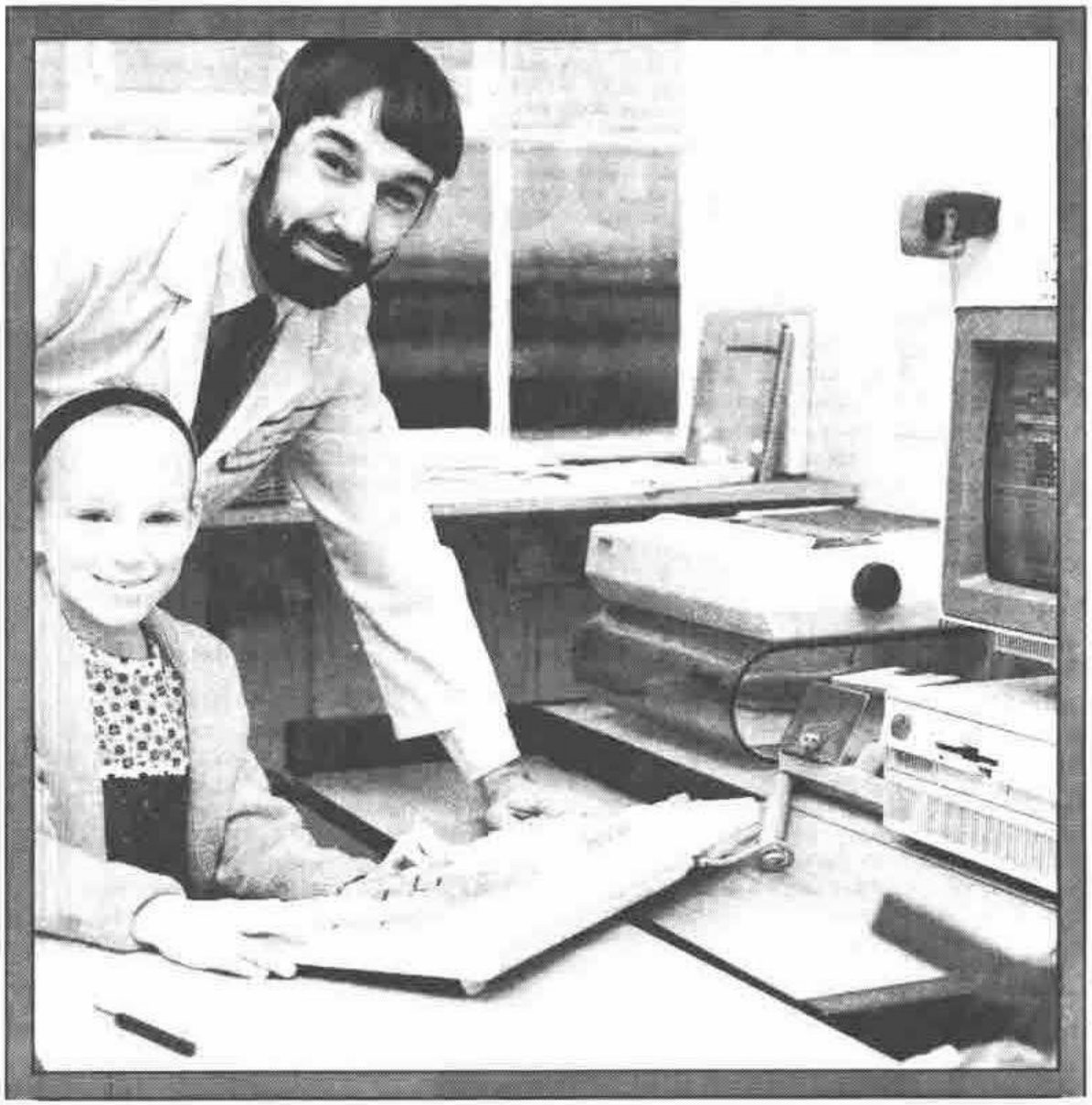 Photograph of a bearded man in a white coat leaning over a smiling girl seated at a PC keyboard