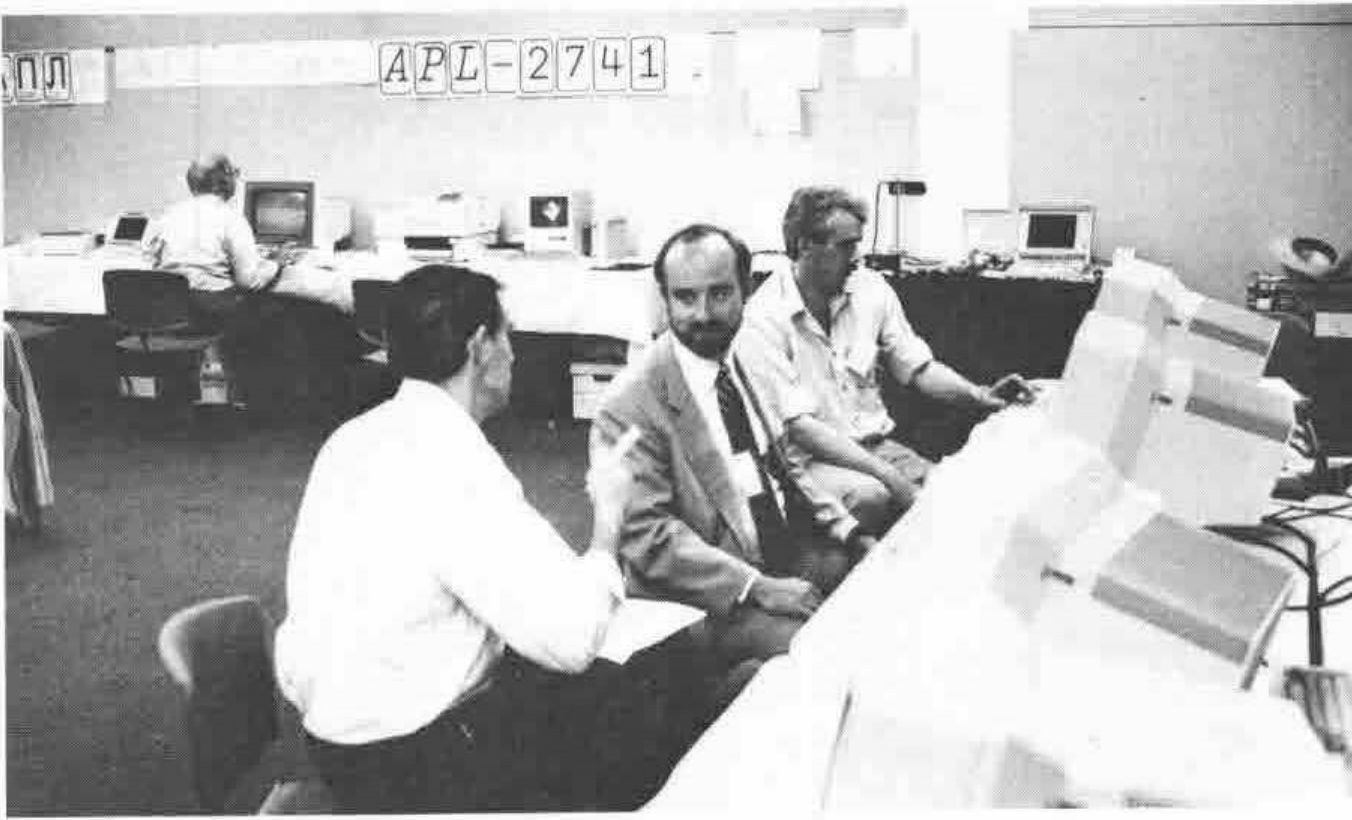 Three men talking among rows of computers, under a banner reading APL-2741