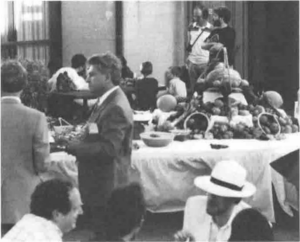 A crowded reception hall with a buffet table piled with fruit; a man in a white hat in the foreground