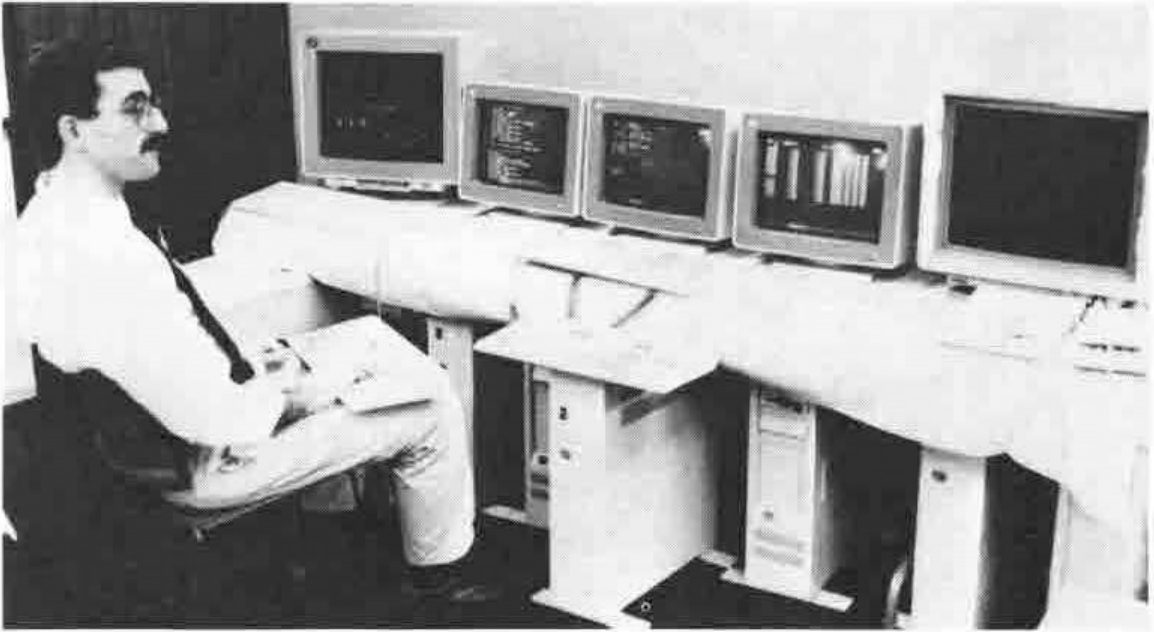 A man seated at a long desk facing a row of five computer monitors