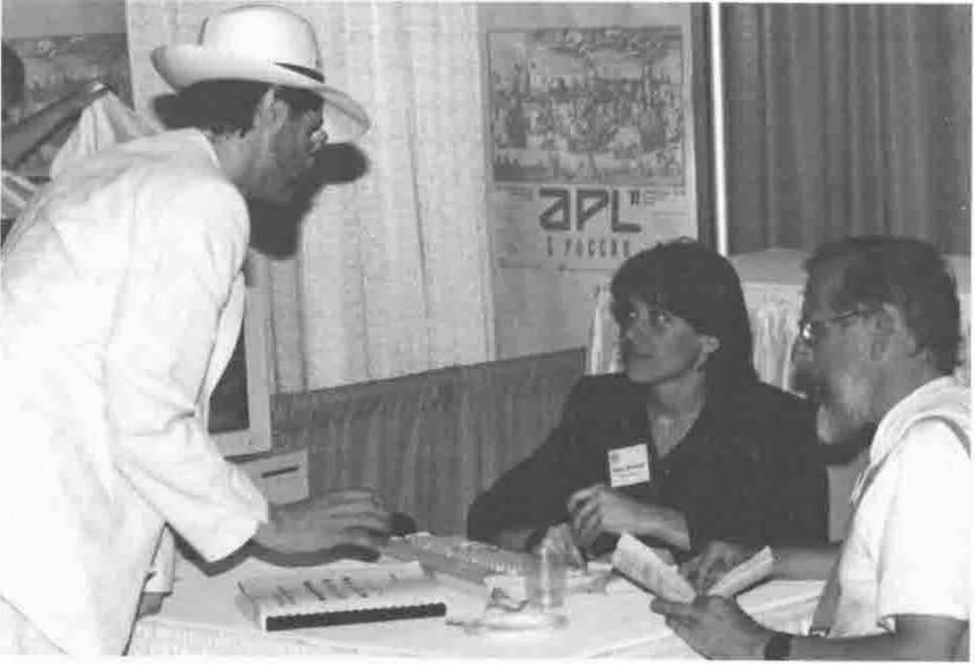 A bearded man in a white hat and jacket leans over a table towards a woman and a bearded man in glasses, seated before an APL poster