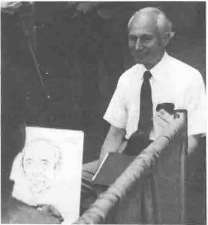 An elderly man in a white shirt and tie at a lectern, beside a pencil portrait of himself