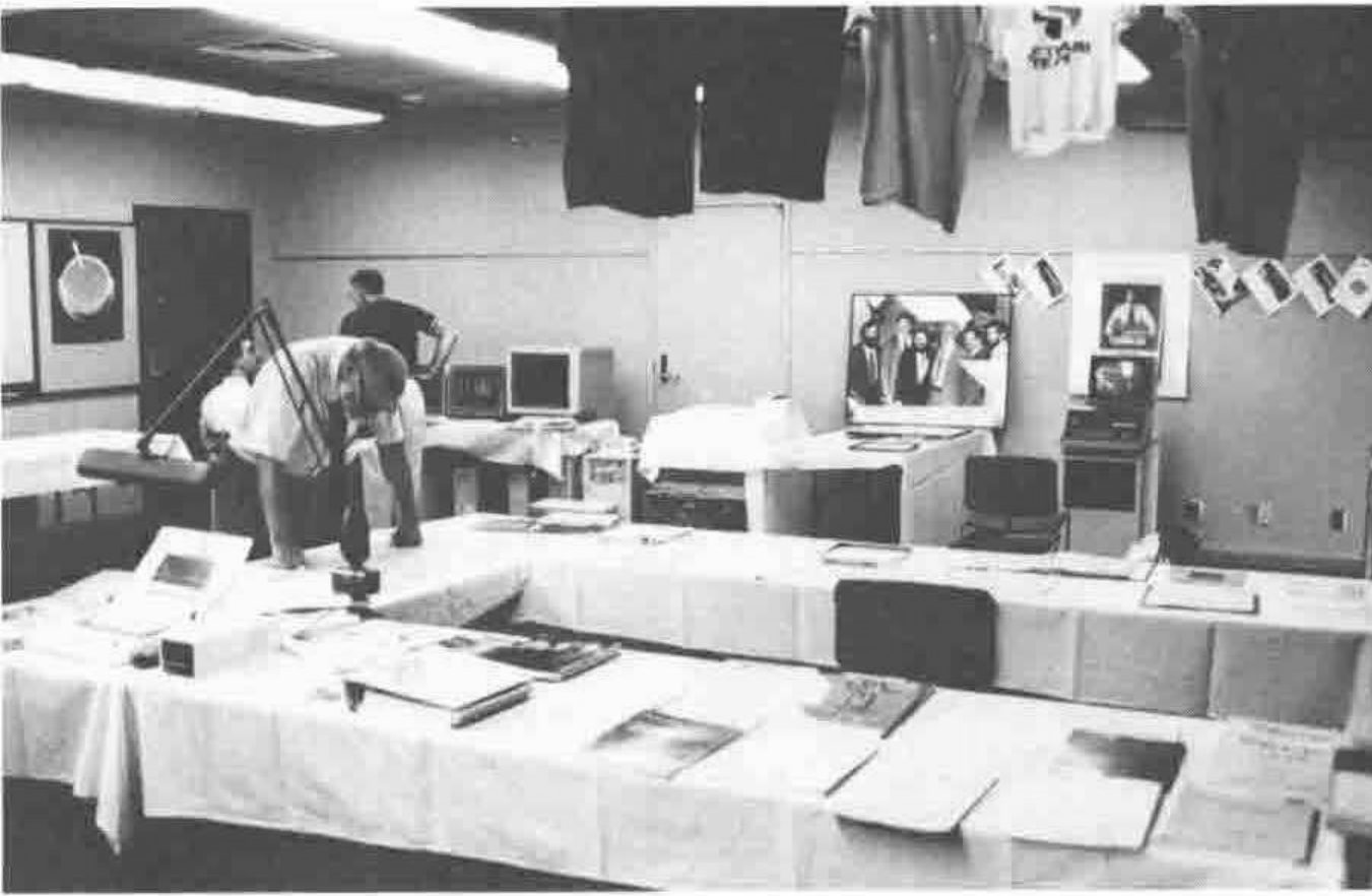 A room with tables of books and papers, early computers, and T-shirts hung from the ceiling; a man leans over one display