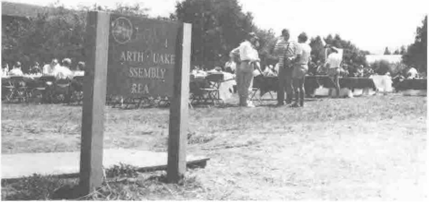 Picnic tables and groups of people on a lawn, behind a wooden sign reading “Earthquake Assembly Area”, partly obscured