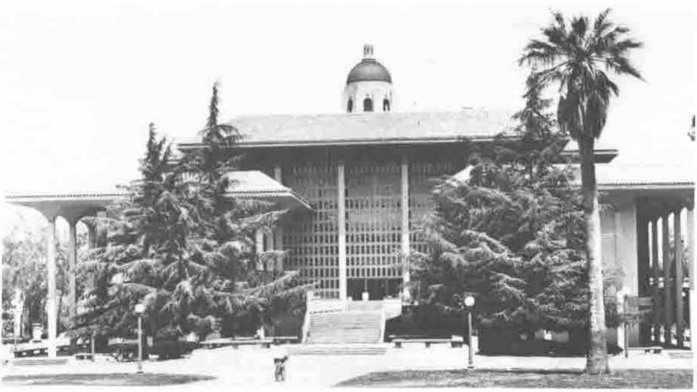 A modern colonnaded library building among trees and a palm, with a domed tower rising behind