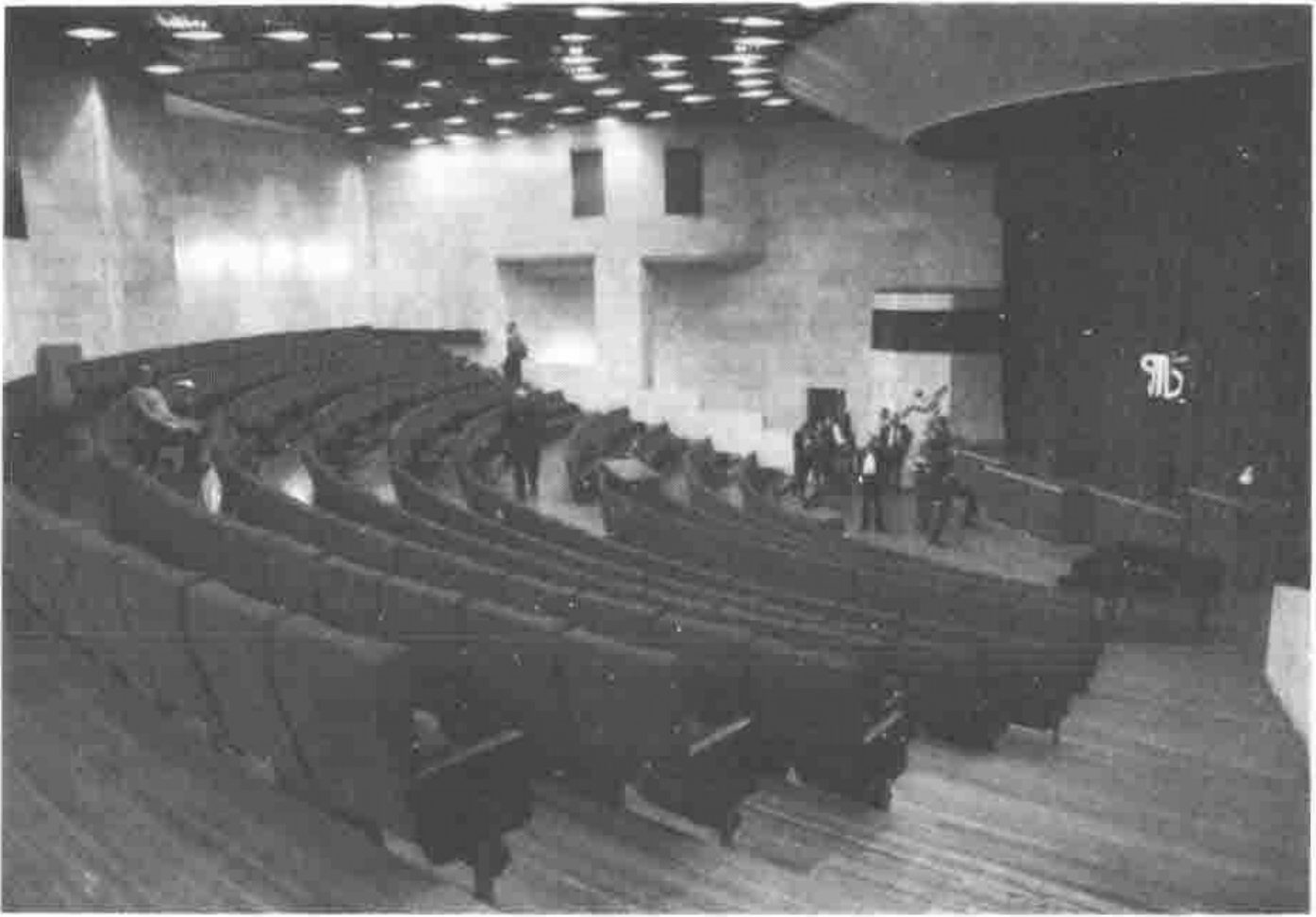 Photograph of a large raked auditorium with curved rows of seats, a few people standing near the stage