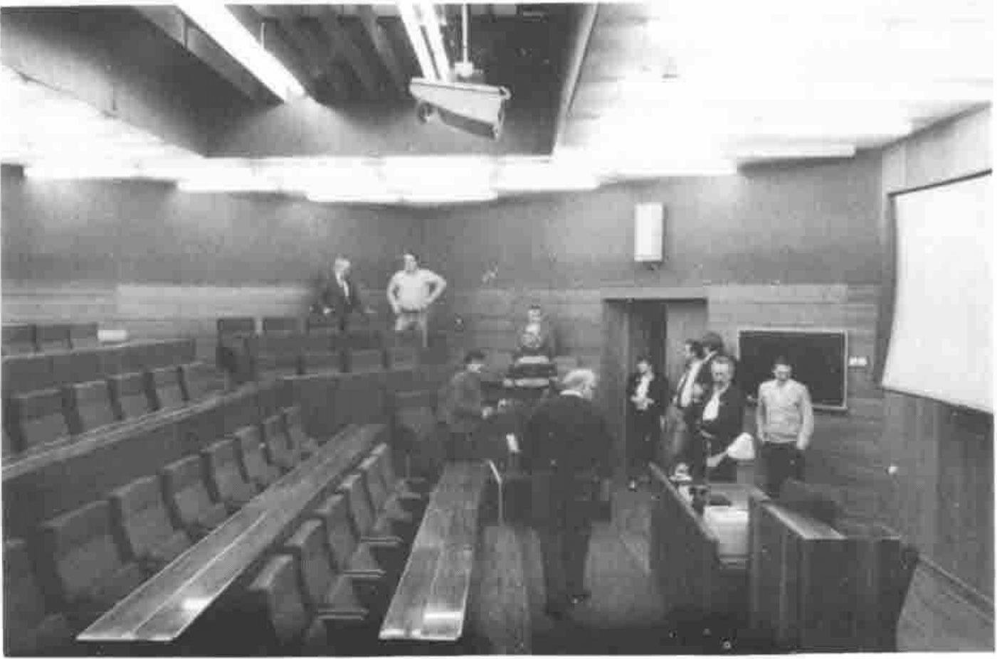 Photograph of a lecture theatre with raked rows of seats and a projection screen, a group of people standing at the front