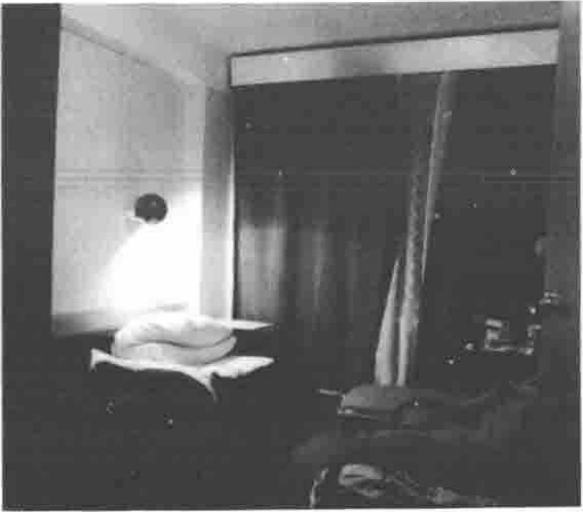 Photograph of a dim single bedroom: a bed with pillows under a wall lamp, curtains and a wardrobe behind