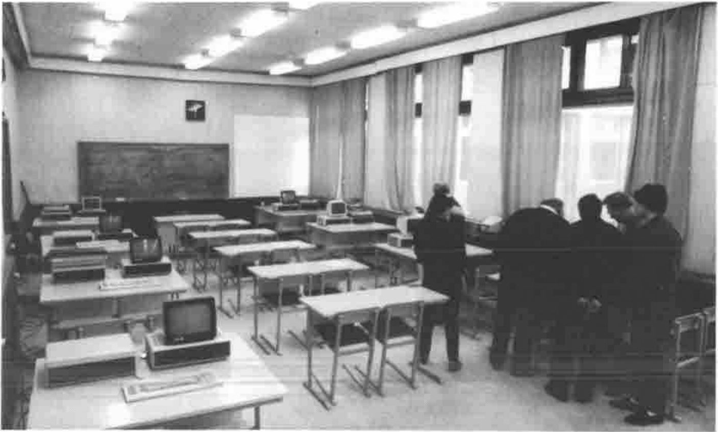 Photograph of a classroom with rows of desks carrying personal computers, a blackboard, and a group of people standing by the windows