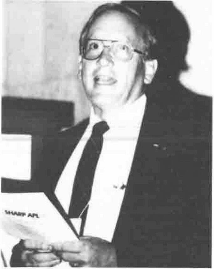 A smiling man in glasses and a dark suit holding open a booklet marked “SHARP APL”
