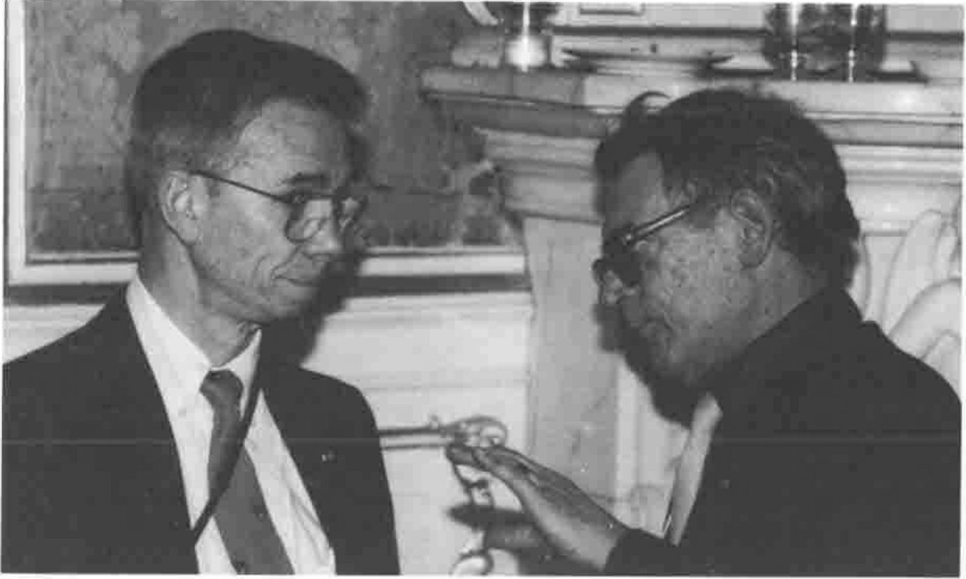 Two men in conversation, one gesturing with a raised hand towards a chain of office worn by the other