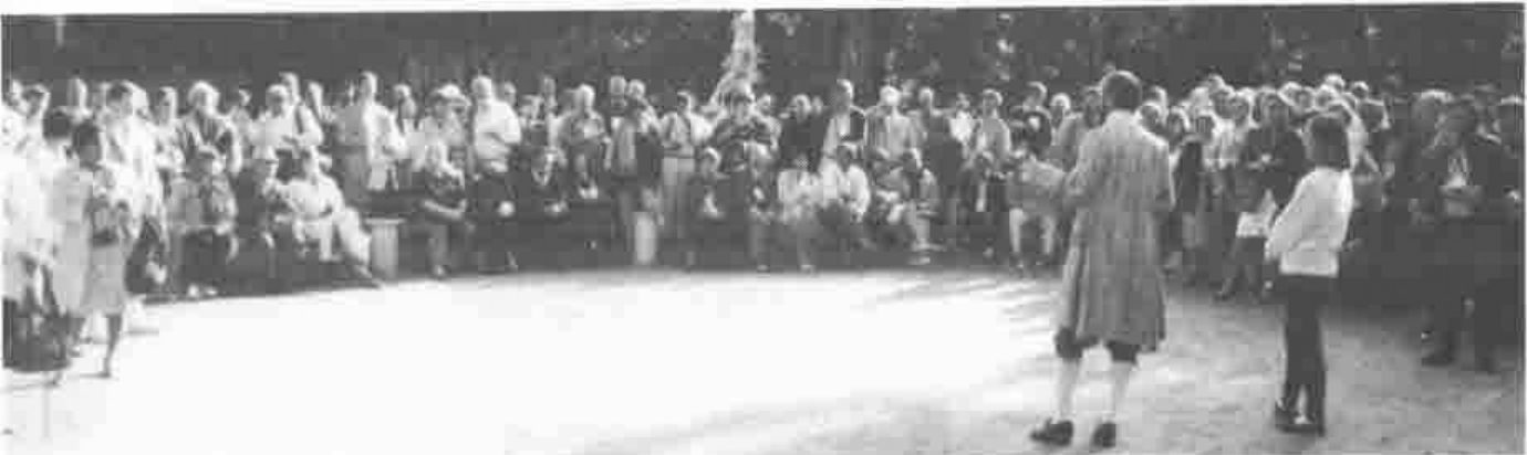 A crowd seated in a semicircle outdoors watching a performer in eighteenth-century costume