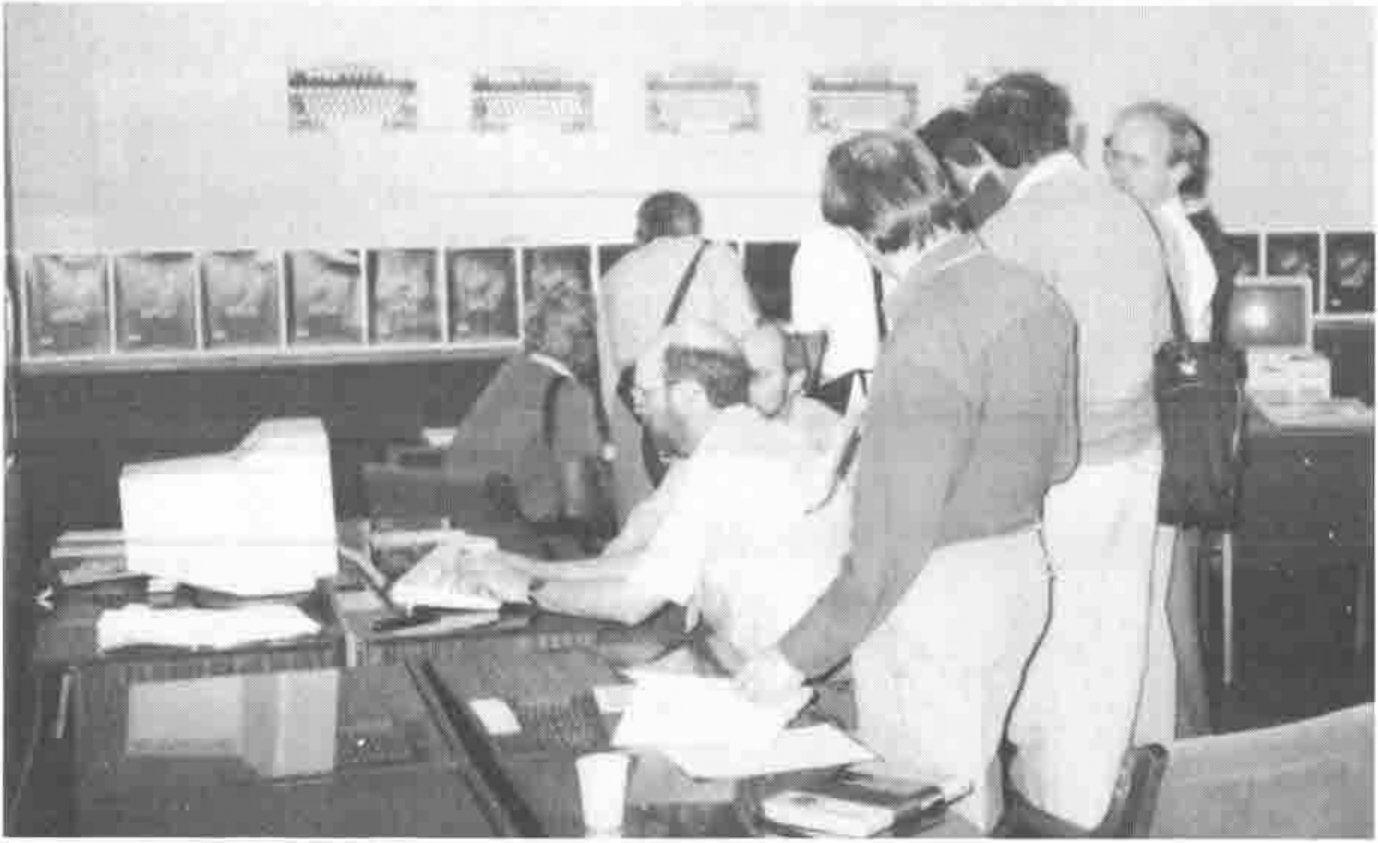 Visitors crowded round a seated man at a computer on an exhibition stand, with a row of posters and keyboard pictures on the wall behind
