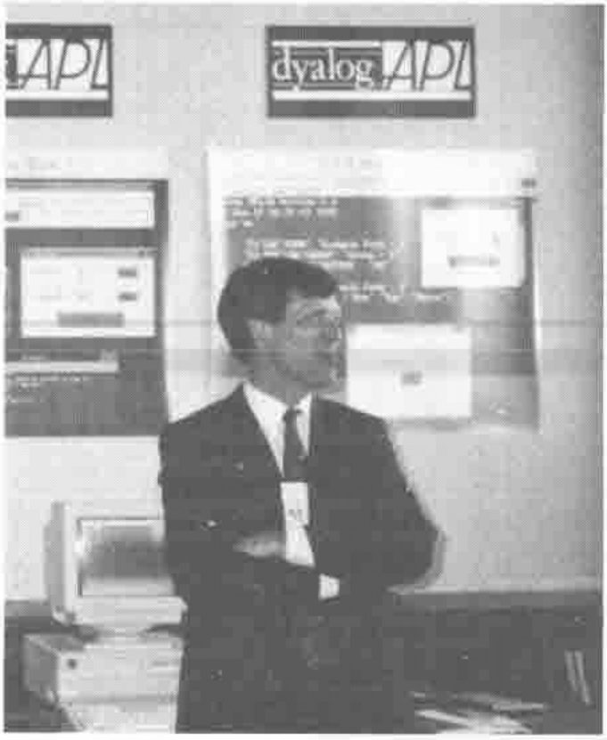 A man in a suit, arms folded, standing in front of a Dyalog APL display stand